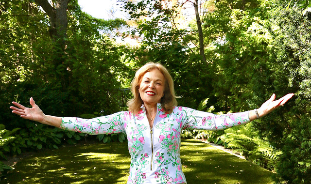 Dr. Elaine Dembe standing joyfully in a garden with arms outstretched